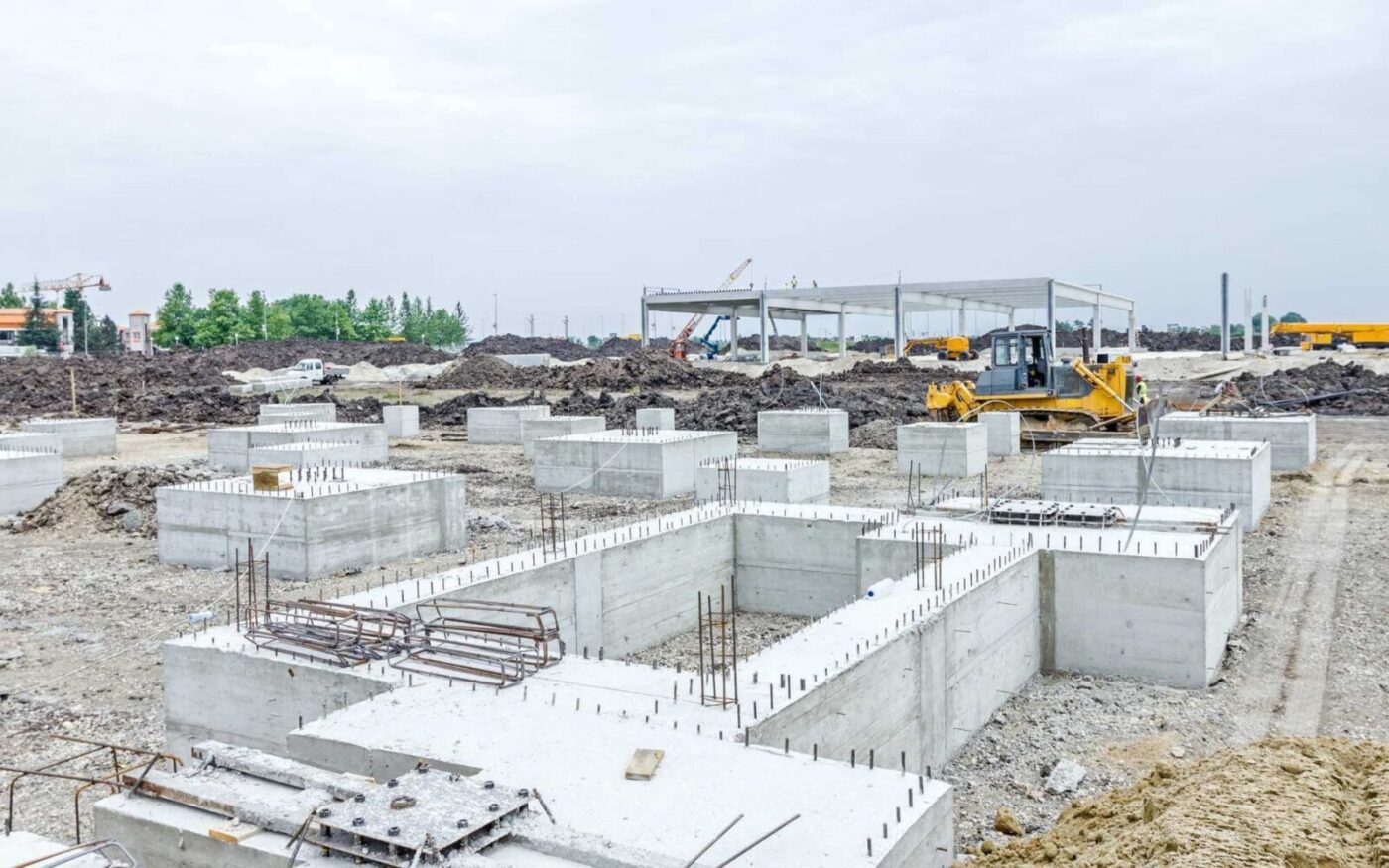 A construction site with concrete foundations laid out on the ground by Naples Concrete Solutions. In the background, construction equipment including a bulldozer and a partially erected structure are visible. The sky is overcast and the surrounding area has piles of dirt and construction materials.