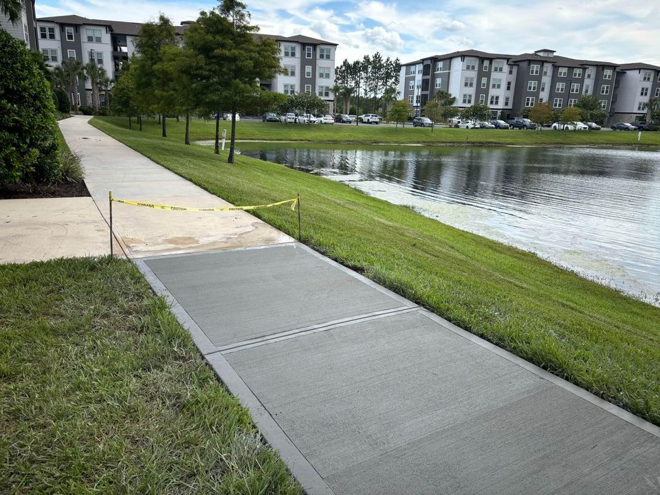 A freshly poured decorative concrete sidewalk, bordered by wooden forms supported by stakes, is curing next to a grassy area and an asphalt road in Naples, FL. Yellow caution tape is attached to the stakes along the sidewalk.