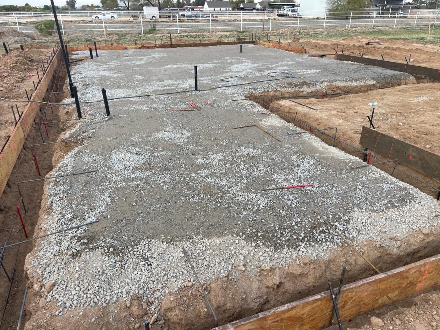 The construction site of the house foundation features wooden formwork and fresh concrete. Several beams and planks support the structure, with metal rebar visible in the concrete. Surrounded by a fence and sandy soil, this concrete project showcases the meticulous work of skilled concrete contractors.