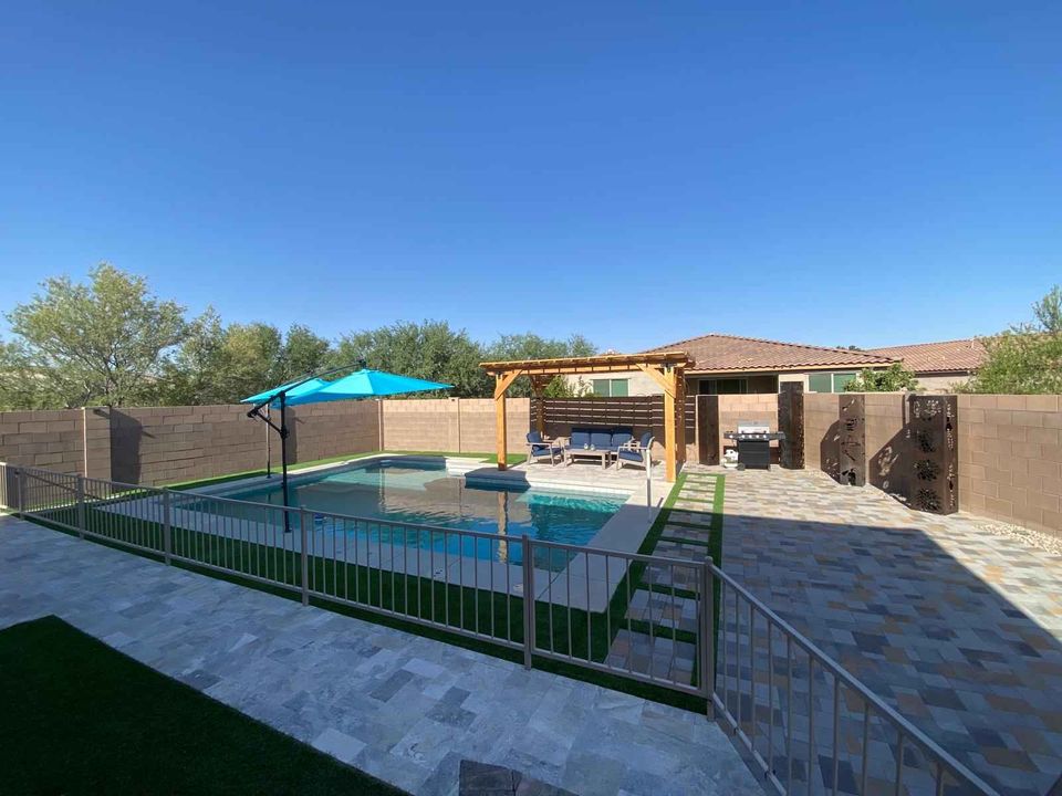 A backyard oasis in Naples, FL, features a swimming pool, embraced by a stone patio crafted with expert paver installation. A metal fence and wooden pergola offer shade with seating underneath. By the pool sits a blue umbrella, all surrounded by a tall brick wall. Majestic trees rise in the distance.