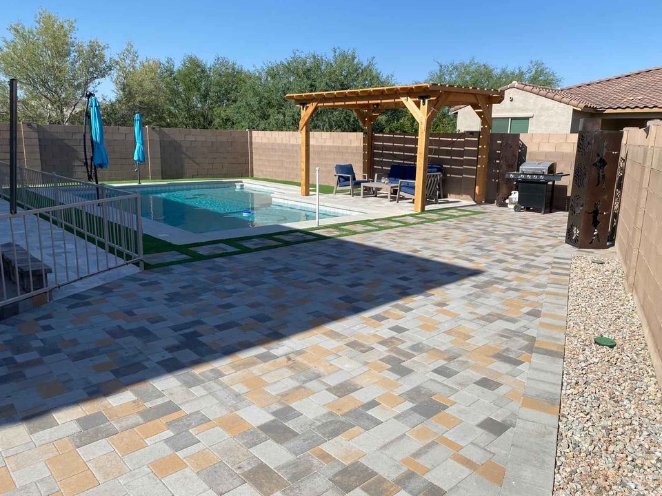 A backyard features a swimming pool with a safety fence, a pergola with seating, and a barbecue grill. The space showcases paver installation with a mix of tile and gravel landscaping. Blue umbrellas provide shade over the patios, while trees flourish in the background.