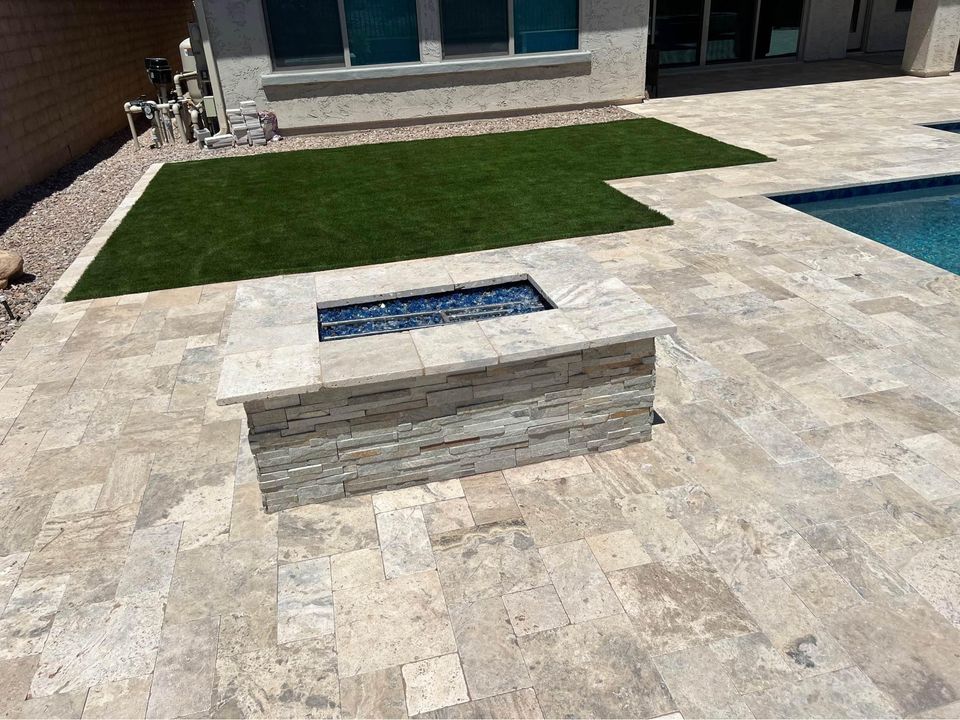 A backyard patio in Naples, FL features a stone firepit set in the center, surrounded by light-colored tiles. Nearby is a rectangular patch of artificial grass and a swimming pool on the right side, showcasing expert paver installation typical of Southwest Florida style.