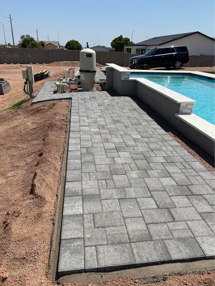 A newly constructed poolside area features gray interlocking pavers, showcasing expert paver installation. In the background, a black SUV is parked near a partially built landscape with exposed dirt and construction materials, characteristic of projects in Southwest Florida.