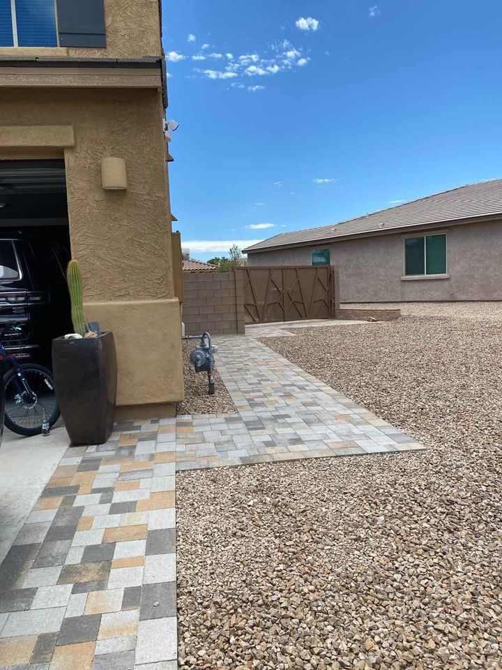 A desert-style backyard features a gravel ground with a gray and brown tile walkway, showcasing expert paver installation. A tall potted cactus thrives near the garage, while the flat-roofed house sits under a clear blue sky.